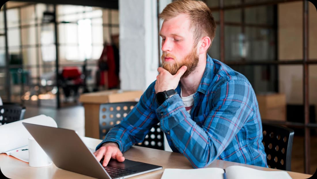 Bearded man in a plaid shirt thinking with one hand on a laptop