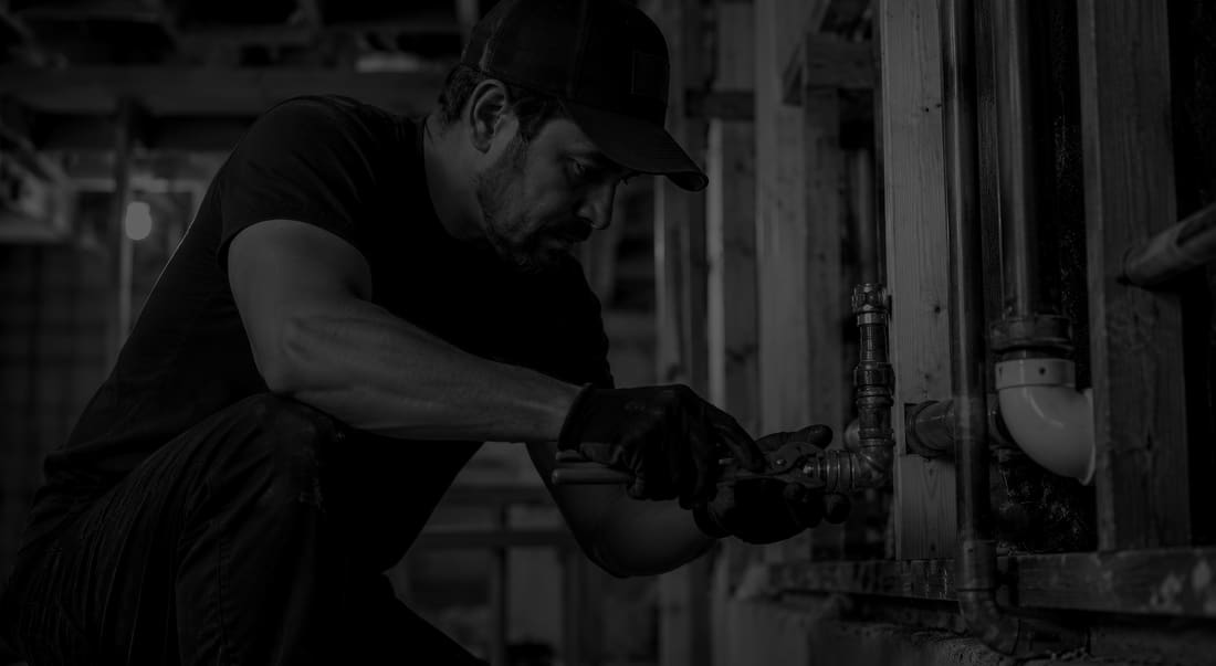 Man in a cap crouching to turn a wrench on pipework in bare framing