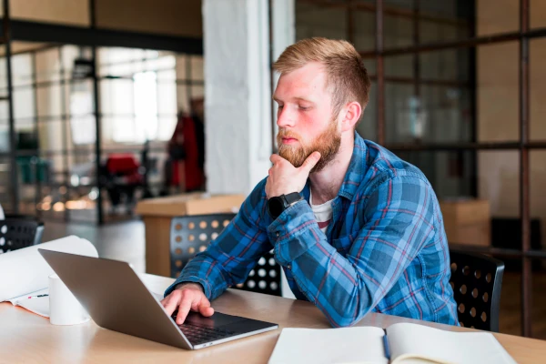 Bearded man at a laptop resting a hand on his chin, looking away