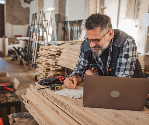 Bearded man in a plaid shirt writing notes beside a laptop on stacked lumber
