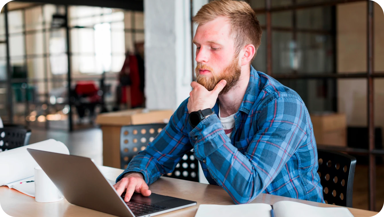 Bearded man in a plaid shirt thinking with one hand on a laptop
