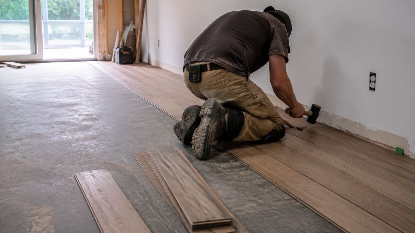 Man kneeling to tap a floor plank into place with a rubber mallet
