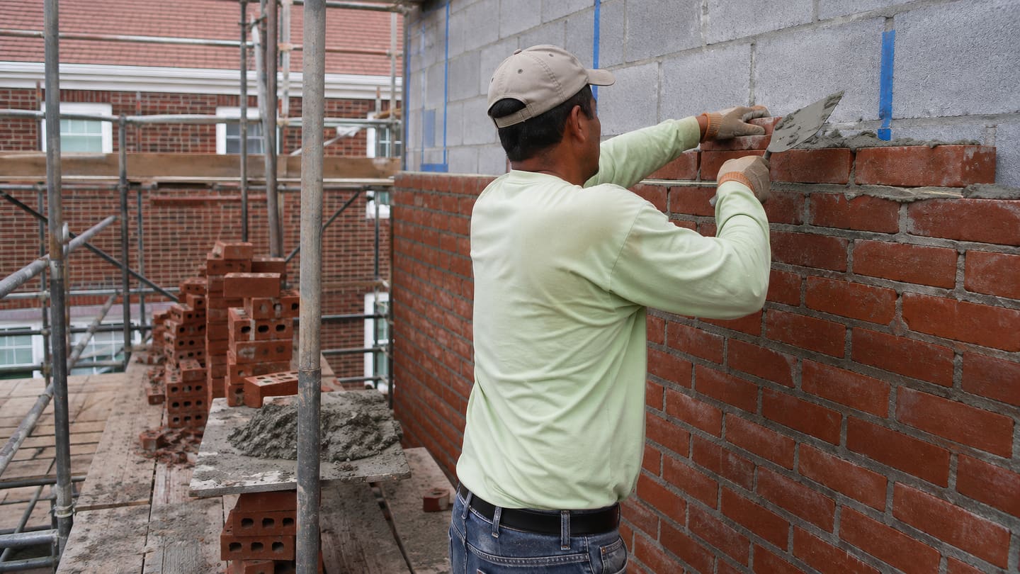 Man on scaffolding spreading mortar with a trowel along a red brick course