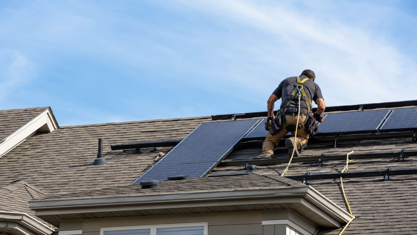 Man in a safety harness setting a solar panel onto roof mounting rails